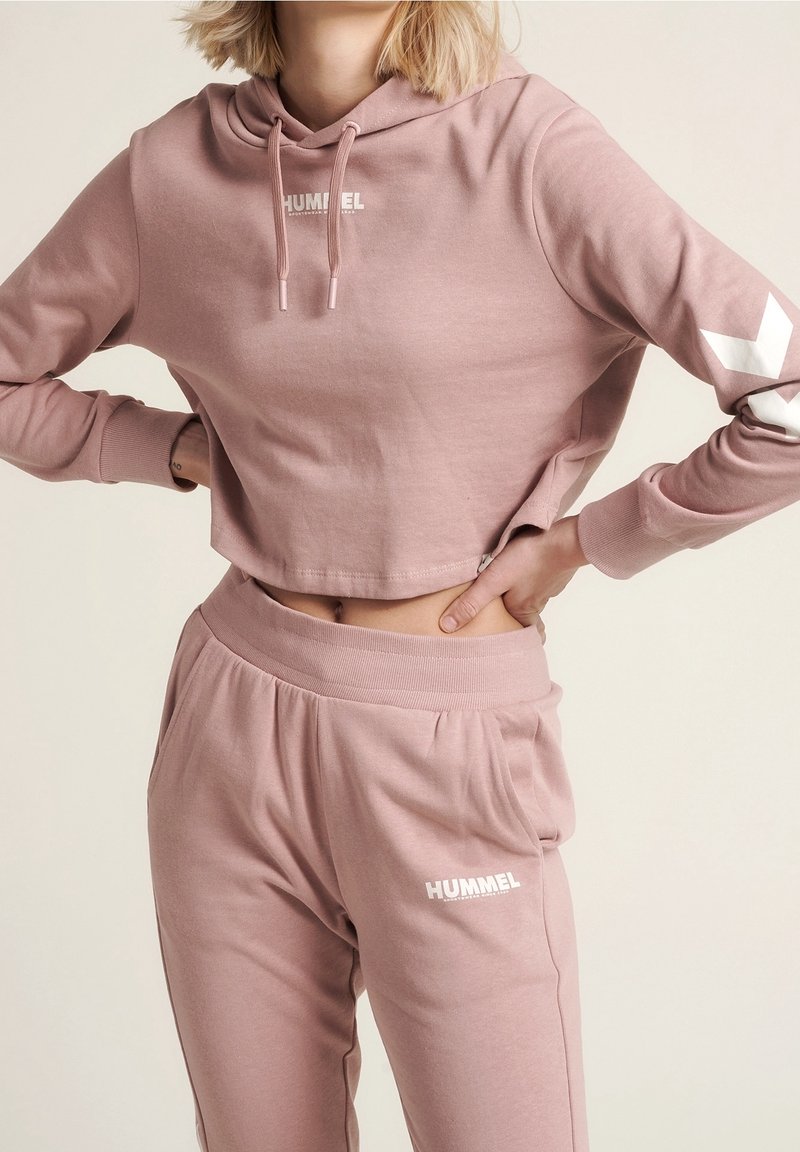 Femme portant un sweat court à capuche rose et un pantalon de survêtement assorti avec logo colibri, mains sur les hanches, fond clair.