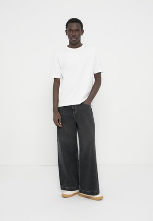 Jonge man die tegen een witte muur staat, gekleed in een effen wit T-shirt, zwarte wijde jeans en witte sneakers met gumzolen.