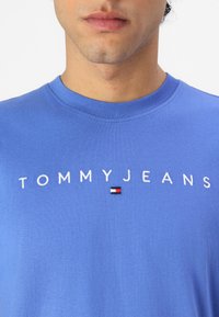 Blå bomulds t-shirt med rund hals, prydet med hvid broderet "TOMMYJEANS" tekst og et lille logo i rød, hvid og blå. Glat tekstur.