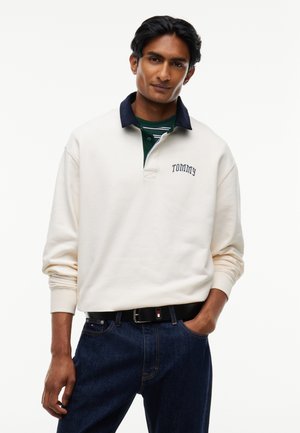 Mand iført cremefarvet langærmet polo med marineblå krave og teksten "Tommy", mørke jeans og sort bælte med logo, poserer med den ene hånd i lommen.