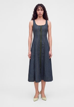 Femme aux cheveux bouclés portant une robe midi en denim foncé sans manches avec une fermeture éclair à l'avant et des chaussures à talons beiges, debout sur un fond uni.