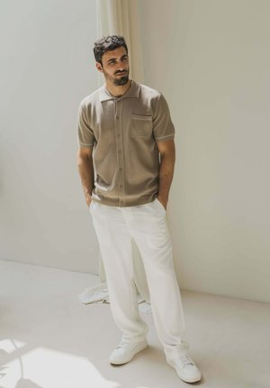 Homme aux cheveux foncés et à la barbe, portant un polo à manches courtes beige à boutons, un pantalon blanc et des baskets blanches, se tenant avec les mains dans les poches.