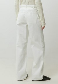 Witte hoge taille jeans met wijde pijpen, voorzien van twee achterzakken en riemlussen. Zachte, gestructureerde stof met een gladde afwerking. Zwarte schoenen zichtbaar.