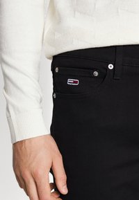 Man wearing black jeans with a small embroidered logo on the front pocket and a white textured long-sleeve sweater.