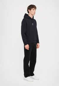 Marineblauer Kapuzenpullover aus einem Baumwoll-Mischgewebe, ausgestattet mit einer Vorderasche und Kordeln. Kombiniert mit schwarzen Hosen und weißen Sneakern.