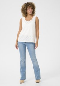 Top bianco senza maniche con design ad occhio, scollatura a girocollo, vestibilità rilassata, abbinato a jeans a zampa d'elefante blu chiaro e scarpe beige.