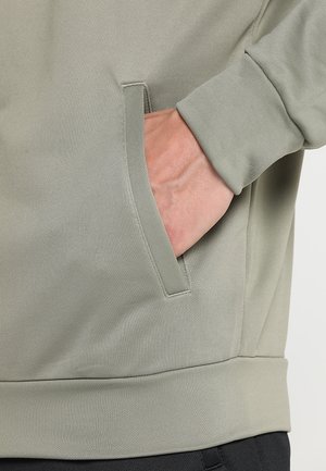 Hand teilweise in die Vordertasche eines hellgrauen Sweatshirts mit langen Ärmeln und geripptem Bündchen und Saum gesteckt.