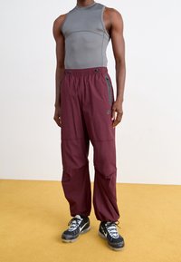 Heren sportoutfit bestaande uit een grijze mouwloze top en bordeauxrode broek met ritszakken, van zachte stof en met een ontspannen pasvorm. Zwarte schoenen met blauwe accenten.