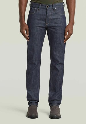 Mann trägt dunkelblaue Jeans mit geradem Bein und oranger Naht, kombiniert mit braunen Knöchelstiefeln, steht vor einem schlichten Hintergrund.