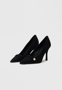 Sorte semskede høye hæler med spiss tå, med et gyllent knappdetalj og en buet design langs forfoten, på en slank hæl.