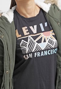 Czarna koszulka z grafiką przedstawiającą góry i słońce, nadrukowaną w różnych pastelowych kolorach. Nałożona pod zieloną kurtkę z futrzanym wykończeniem.