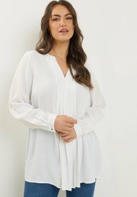 Blusa bianca a maniche lunghe con scollo a V, fronte plissettato e polsini con bottoni, abbinata a jeans blu su uno sfondo neutro.