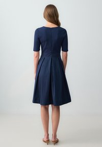 Navy blauwe jurk met korte mouwen, ronde halslijn, getailleerde bovenkant en een uitlopende, geplisseerde rok. Ritssluiting aan de achterkant. Soepele textuur.