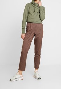 Persona che indossa una felpa verde oliva, pantaloni slim-fit a quadri marroni e sneakers bianche con particolari rosa, in piedi davanti a uno sfondo semplice.