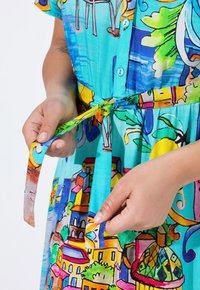 Bunt gemustertes Baumwollkleid mit blauer Basis, das lebendige Motive von Häusern, Pflanzen und abstrakten Formen zeigt. Mit einem gebundenen Taillendetail.