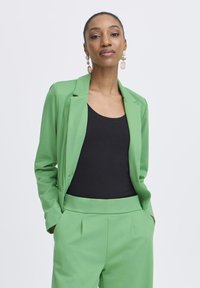 Vrouw met naar achteren gebonden haar, gekleed in een groen colbert en broek, een zwart topje, en hangende oorbellen, staand met handen in de zakken.