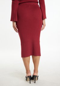 Brede midi-jurk in diep rood, geribbelde textuur, figuurvormende silhouet, met een licht asymmetrische zoom. Draag het met zwarte hoge muiltjes.