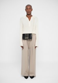 Borsa crossbody nera con design a borchie e lettering dorato, abbinata a un top in maglia crema e pantaloni beige a gamba larga. Tacchi a punta neri.