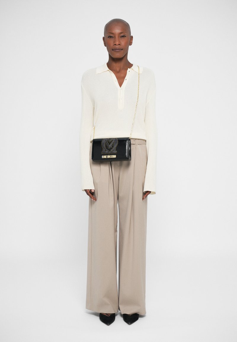 Borsa crossbody nera con design a borchie e lettering dorato, abbinata a un top in maglia crema e pantaloni beige a gamba larga. Tacchi a punta neri.