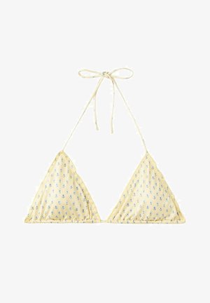 Femme en bikini jaune à fleurs ajustant le bas près d’une eau claire et peu profonde avec des collines rocheuses sous un ciel bleu, portant des lunettes de soleil noires.