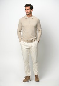 Beige Langarmpolo mit Reißverschlusskragen, kombiniert mit beigefarbenen Hosen und braunen Schnürsneakern auf einem einfarbigen Hintergrund.