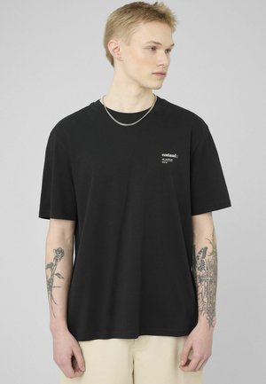 Jeune personne aux cheveux blonds courts portant un t-shirt noir avec un petit texte blanc et un collier chaîne, montrant des tatouages sur les deux avant-bras.