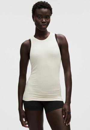 Schwarze Frau steht, trägt ein ärmelloses weißes Top über einem schwarzen Sport-BH und schwarzen, eng anliegenden Shorts, vor einem schlichten hellen Hintergrund.