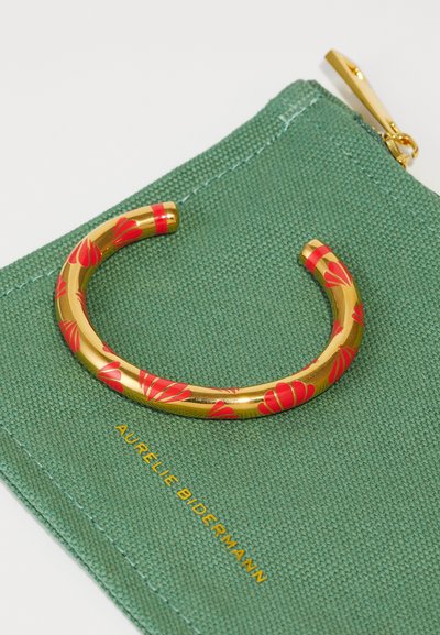 Brazalete de muñeca de oro con patrón floral rojo, descansando sobre un bolso verde texturizado con el texto dorado "AURELIE BIDERMANN."