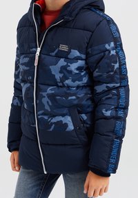 Junge trägt eine marineblaue Camouflage-Puffermantel mit Kapuze, Frontreißverschluss, Seitentaschen und Textdetails auf dem Ärmel und der Brust.