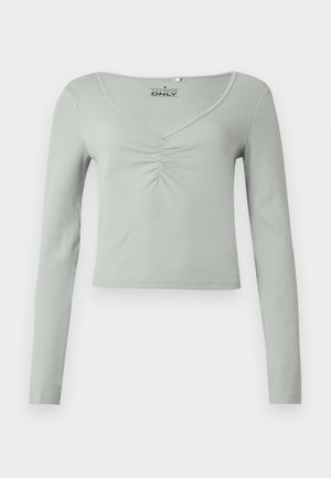 Šviesiai žalios spalvos ilgomis rankovėmis viršus iš gofruotos medžiagos, su V formos iškirpte ir centrinės priekio dalies surinkimu. Trumpintas modelis.