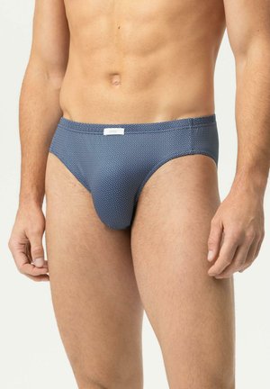 Mann trägt blaue gemusterte Unterhosen und steht vor einem schlichten hellen Hintergrund, der den Oberkörper und die oberen Beine zeigt.