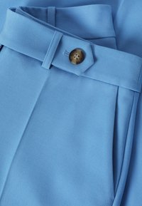 Pantalon en tissu bleu clair avec une texture lisse, doté d'un bouton marron et de plis soigneusement pliés sur la ceinture pour un look ajusté.