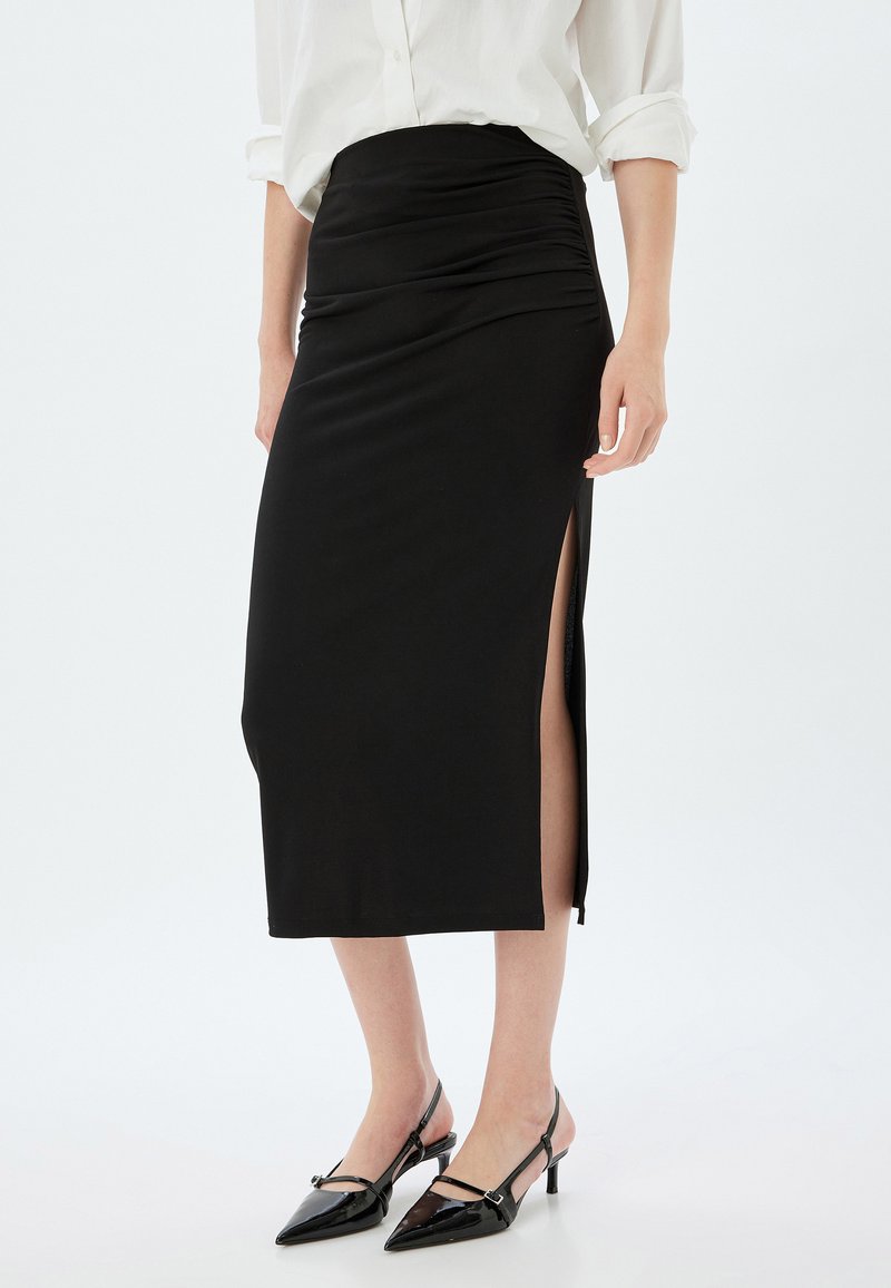 Musta midi-hame, jossa on korkea vyötärö, rypytetty tekstuuri ja sivureikä, yhdistettynä mustiin kärjellisiin slingback-korkokenkiin.