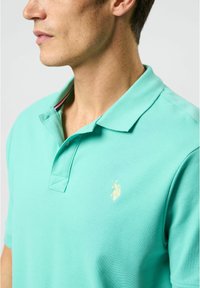 Ljus turkos polo-tröja i texturerad tyg med krage, två knappar och en liten, broderad logotyp i guld på bröstet.