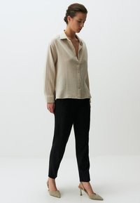 Lässiges beige strukturiertes Hemd mit Knöpfen, kombiniert mit maßgeschneiderten schwarzen Hosen und spitzen beigen Absatzschuhen, die einen subtilen, eleganten Look präsentieren.