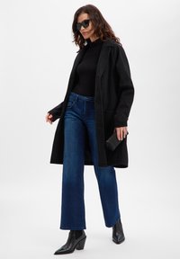 Long manteau noir porté sur un haut noir ajusté, associé à un jean bleu à jambes larges et des bottines noires à bout pointu. Accessoires comprenant des lunettes de soleil foncées.
