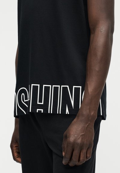 Κοντινό πλάνο του κορμού και του χεριού ενός ατόμου που φοράει μαύρο πουκάμισο με μεγάλα λευκά γράμματα με περίγραμμα κοντά στο στρίφωμα και μαύρο παντελόνι.