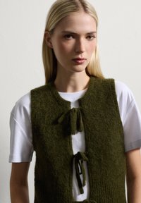 Donkergroene gebreide vest met een ronde halslijn, met twee voorbinders en een gestructureerde afwerking, gedragen over een wit shirt.