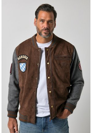 Mand med kort mørkt hår og skæg iført brun og grå varsity jakke med Bavaria-mærke, hvid T-shirt og blå jeans står mod en ensfarvet baggrund.