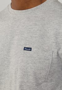 Close-up van een lichtgrijs gemêleerd T-shirt met een klein marineblauw label met de tekst "Façonnable" dat boven de borstzak is genaaid.