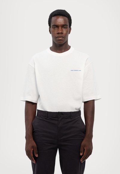 White cotton t-shirt with a round neck and short sleeves, featuring blue text on the chest. Worn with dark trousers.