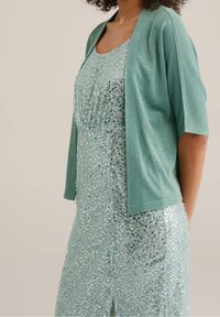 Svetle tyrkysový kardigan přes třpytivé aqua šaty, zdobené flitry, s kulatým výstřihem a krátkými rukávy. Šaty mají boční rozparek.