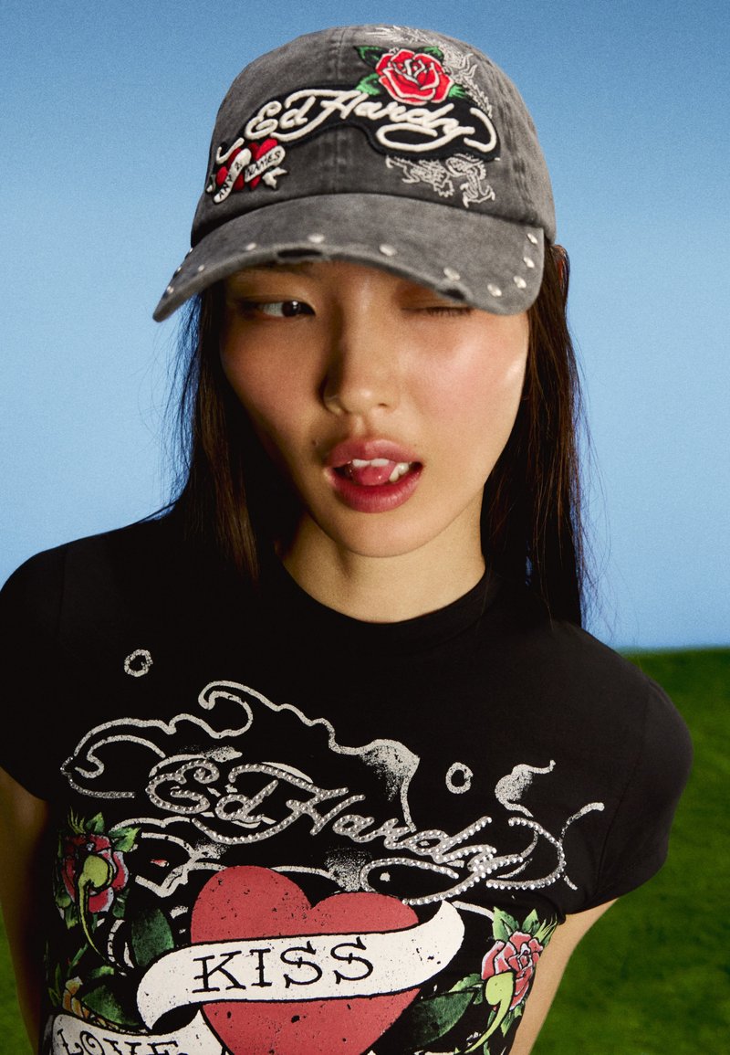 Jeune femme portant une casquette Ed Hardy usée et un t-shirt noir avec un cœur rouge et le texte « KISS », faisant un clin d'œil avec la langue légèrement sortie devant un ciel bleu.