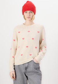 Pull beige à motifs de cœurs roses, réalisé en tissu doux ; il a un col rond et des poignets côtelés, associé à un pantalon gris.