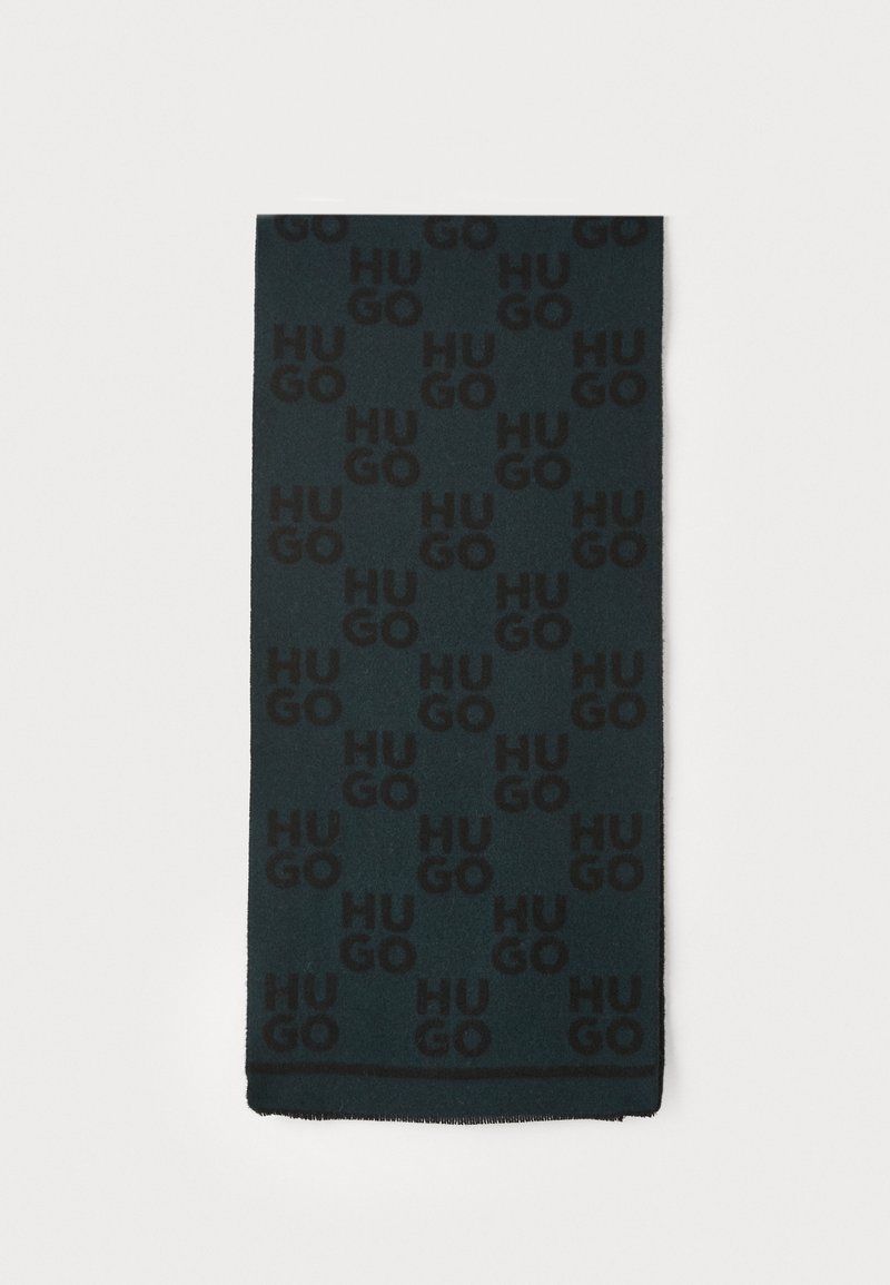 Sort tørklæde med gentagende "HUGO" logo mønster på en mørkegrøn baggrund. Materialet virker blødt med en frynset kant. Rektangulær form.