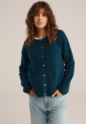 Vrouw met golvend bruin haar draagt een donkerblauwe knopentrui en lichtblauwe spijkerbroek, staand met haar handen in de zakken tegen een effen achtergrond.