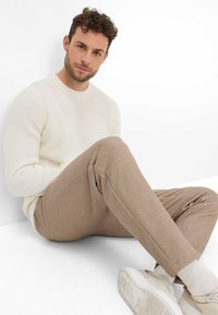 Pull en maille texturée crème, pantalon en carreaux beige, et baskets claires. Modèle assis contre un fond blanc, adoptant une posture détendue.