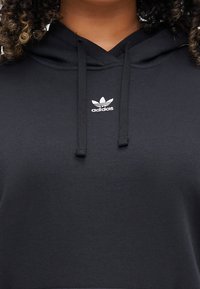 Černá mikina s kapucí vyrobená z měkkého materiálu, s výstrhem do V, šňůrkou a malým bílým logem Adidas na hrudi.