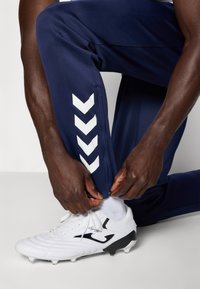 Osoba tamnije puti kleči i namješta patentni zatvarač na mornarskoplavim sportskim hlačama blizu gležnja, noseći bijele kopačke s crnim detaljima.