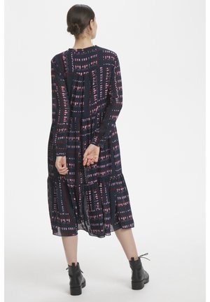 Robe noire avec imprimé graphique en rose et blanc, jupe à volants, manches longues et col rond. Associée avec des bottines noires.
